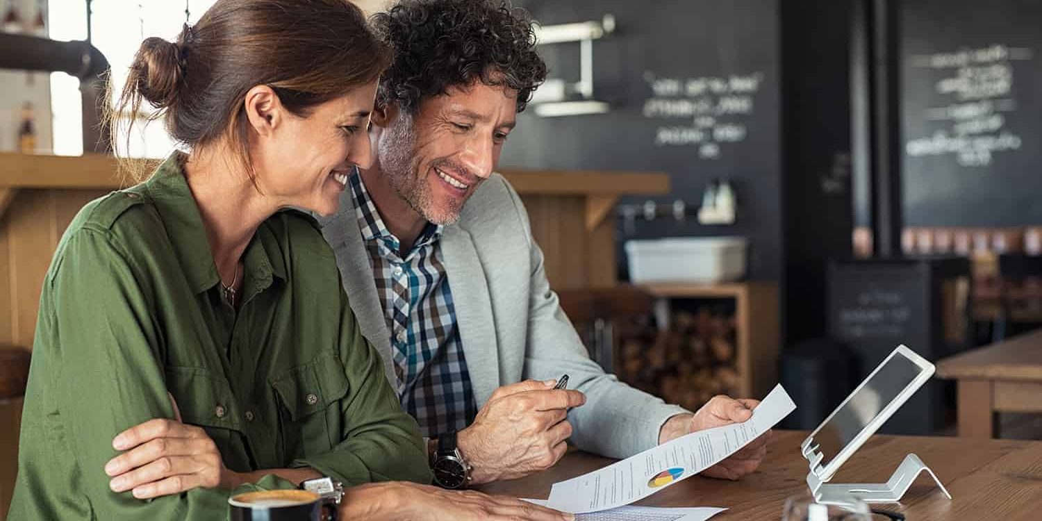 Portrait of mature business man and casual businesswoman sitting in cafe and discussing sales graph. Group of two middle aged coworkers working comparing forecasting graphics. Happy businessman reading report with his business partner.