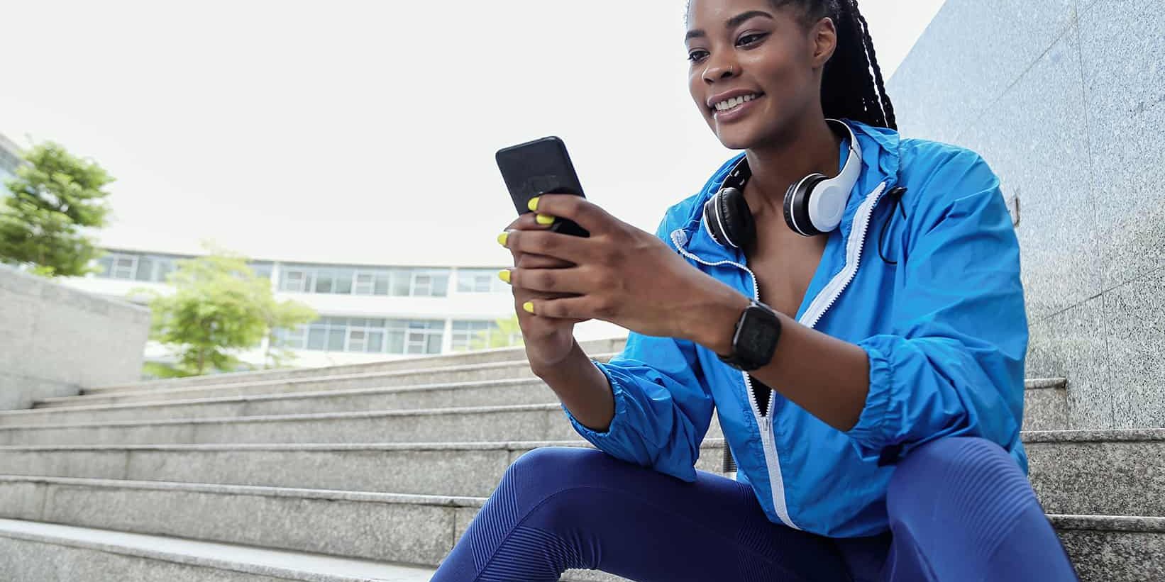 Pretty young smiling sportswoman sitting on steps and reading text messages from friends on smartphone